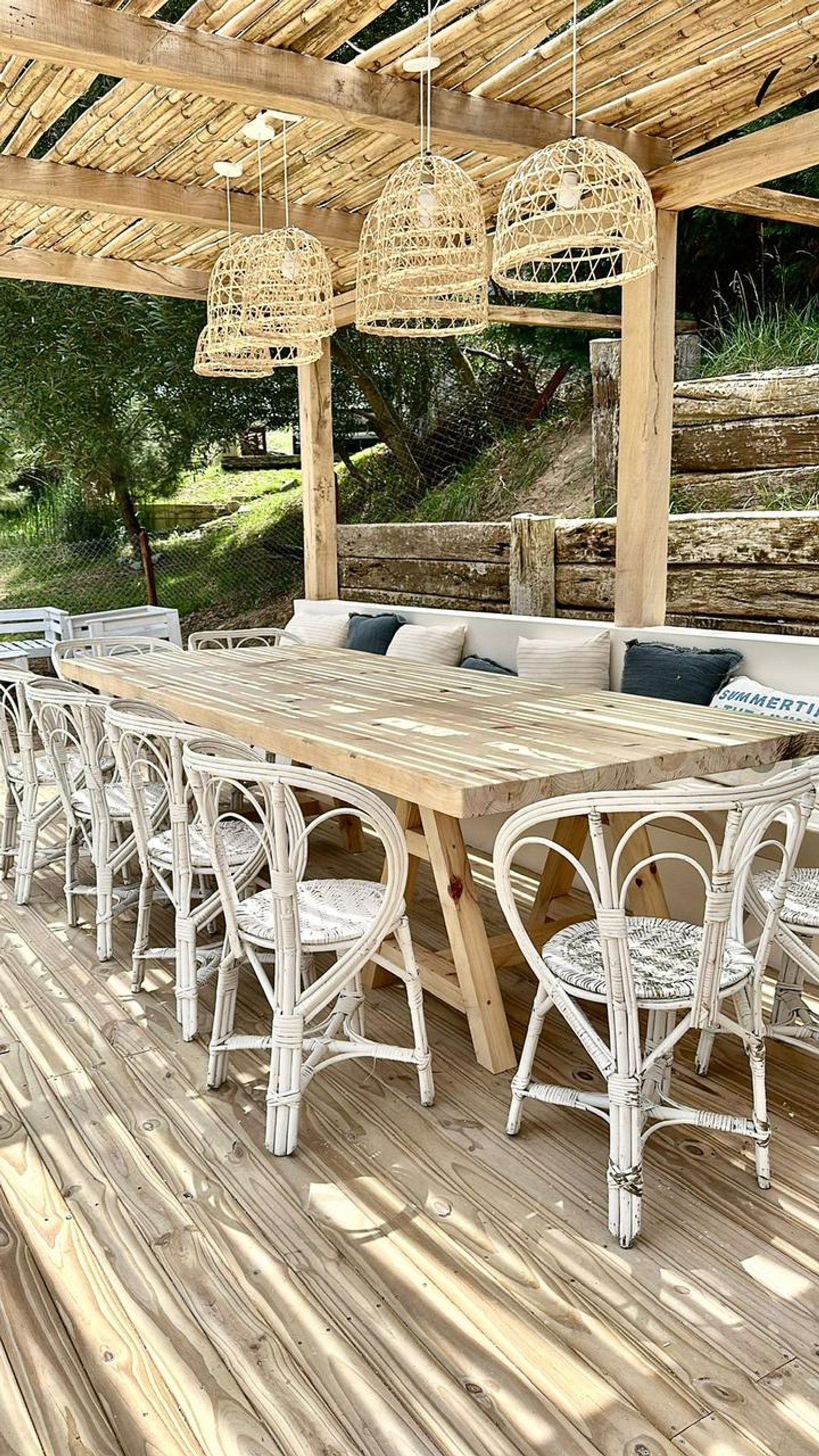 Comedor exterior con mesa rústica de madera y sillas de ratán blancas — proyecto CE, por arquitectura.JD, en Pinamar